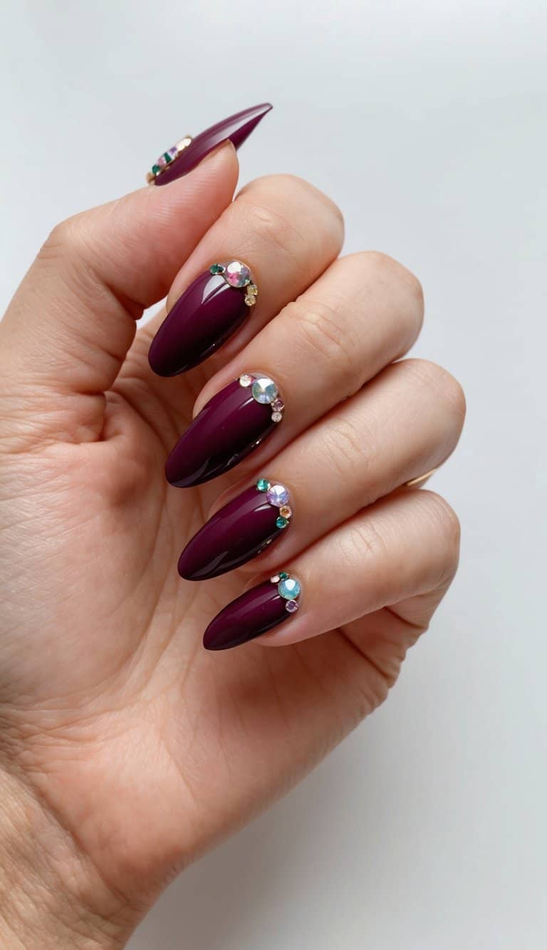 A hand showcasing glossy maroon nail art with sparkling rhinestones and gold embellishments near the cuticles, creating a glamorous and sophisticated design.