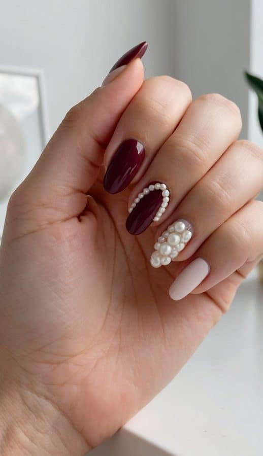 A stylish maroon nail design featuring glossy burgundy, soft nude nails, and pearl embellishments, offering a chic and sophisticated look.