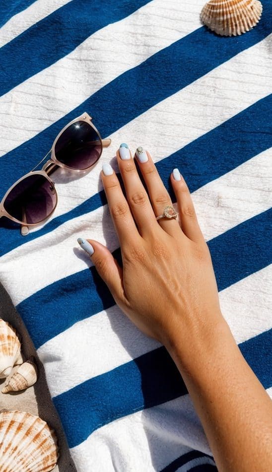 Elegant summer nail design featuring pastel blue and white nails with metallic details, resting on a navy-striped beach towel.