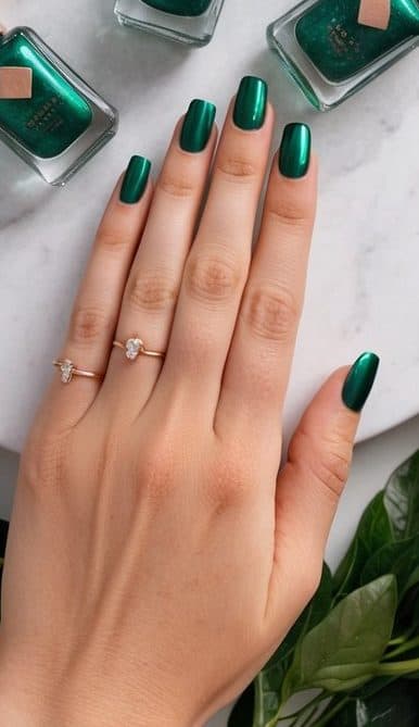 Emerald green nail polish bottles with rose gold foil accents arranged on a marble surface, surrounded by lush greenery and gold accents