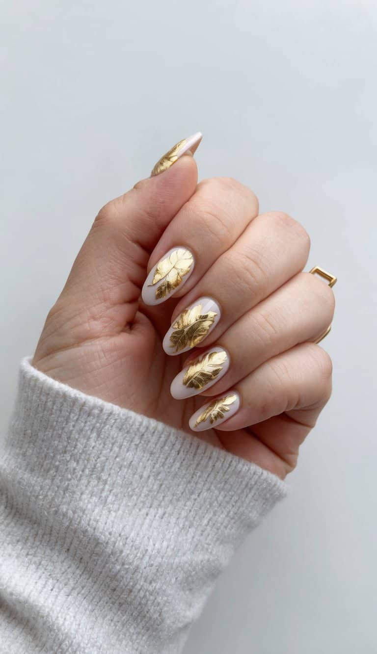 A stylish set of almond-shaped nails with a translucent white base and striking gold leaf accents, creating a modern and formal nail art look.