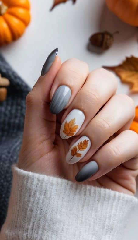 A hand showcasing fall nail art with gray ombre polish and white nails adorned with orange autumn leaves, creating a cozy and artistic seasonal vibe.