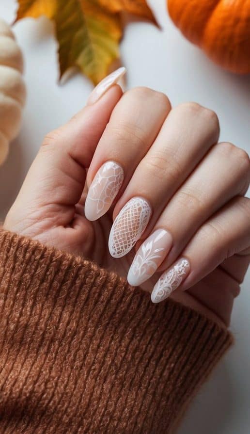 A soft autumn-inspired manicure featuring nude nails with white lace designs, styled with a cozy sweater and fall leaves.