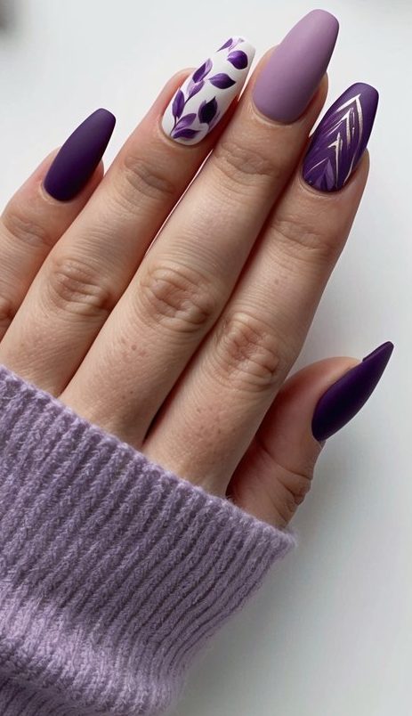 Matte purple nails featuring a soft lavender base, delicate hand-painted purple leaves, and a striking metallic chevron accent design.