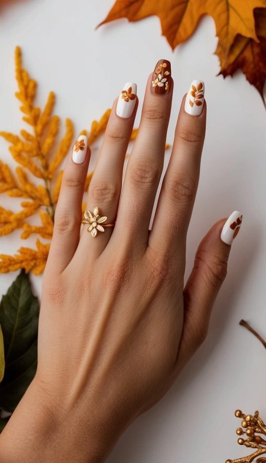 Thanksgiving nails featuring white and terracotta shades with embossed amber flowers and fall leaf details, surrounded by pumpkins and autumn décor.