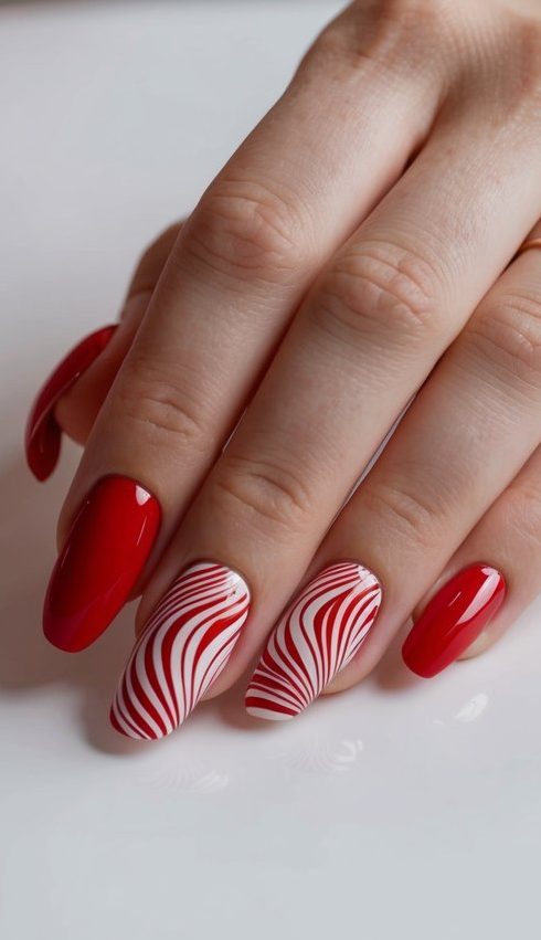 Bright red nails with glossy finish, paired with two accent nails showcasing red and white swirl patterns for a festive and striking manicure.