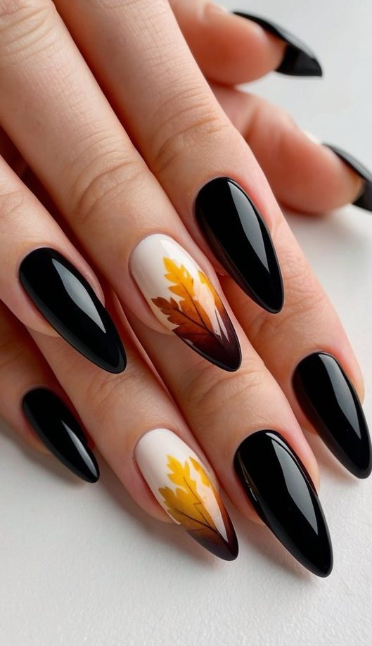 Pointed nails featuring glossy black polish with two accent nails displaying yellow-orange leaves blending into a black ombré on a soft beige base.
