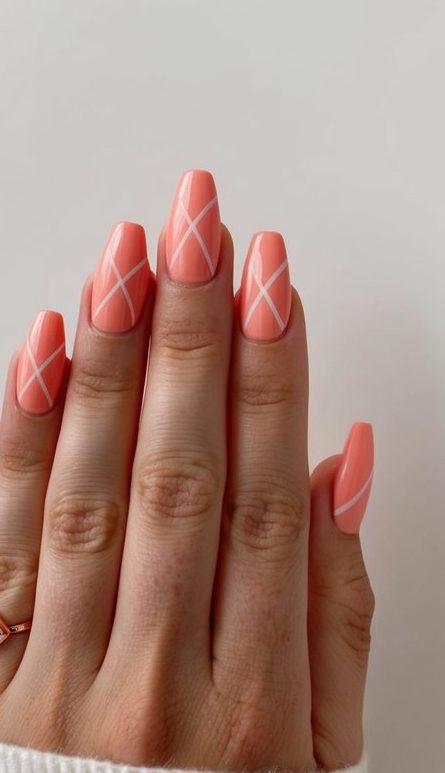 Hand with almond-shaped coral nails featuring white diagonal crisscross pattern emerges from white ribbed sweater sleeve against neutral background, adorned with small gemstone ring.