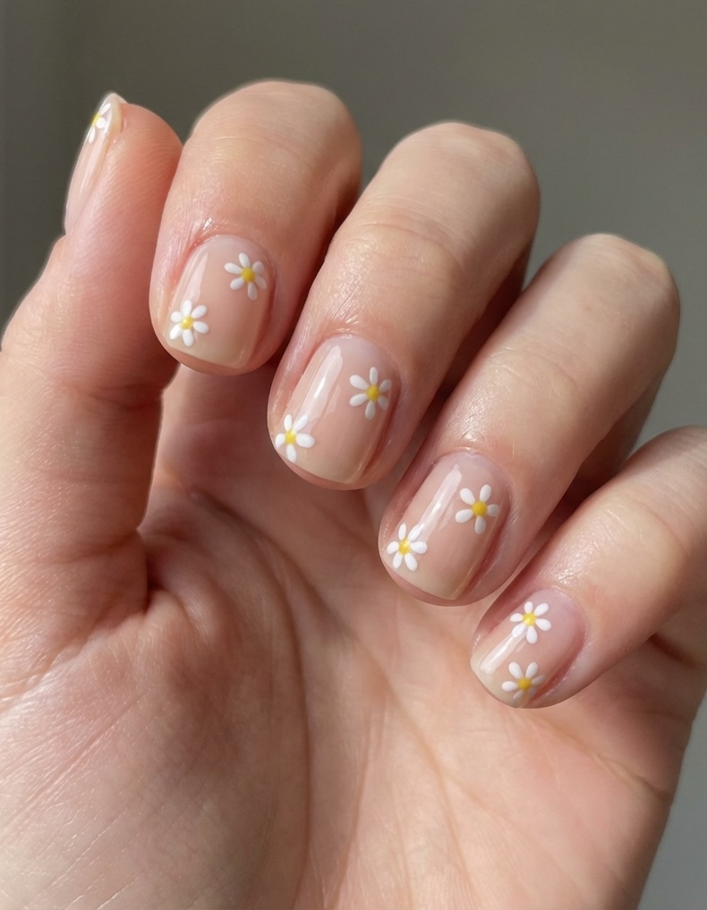 A close-up of a hand with a soft nude manicure featuring a short nail art idea with tiny white daisy flowers and yellow centers on each nail.