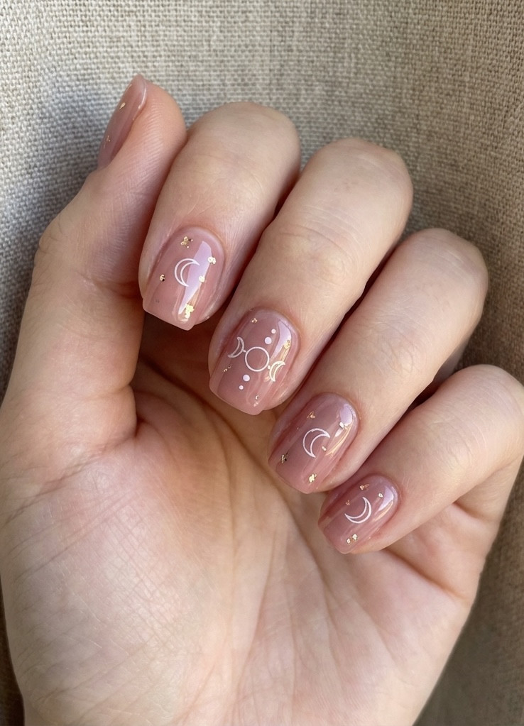 A hand with neatly squared nails shows a dusty rose nail art idea using white celestial symbols and gold flakes on a pink-beige glossy base.