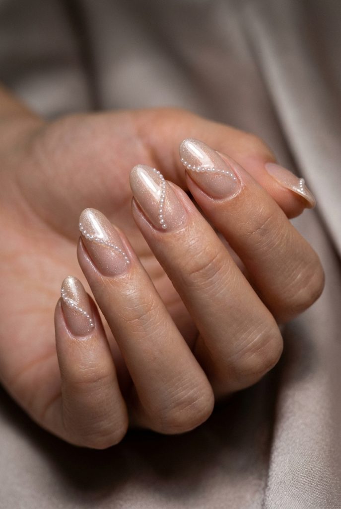 A delicate hand holds still as curved trails of micro-pearls sweep over sheer champagne nails, showcasing an intricate pearl nail art idea under soft lighting.