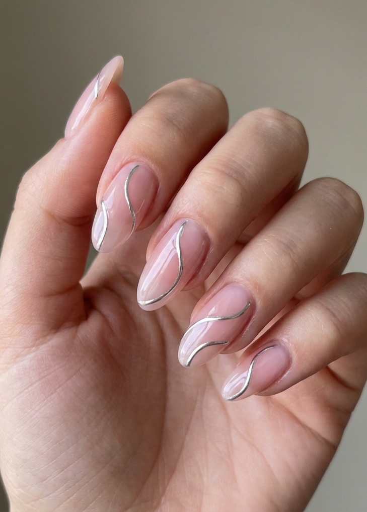 Close-up of modern glazed nail art idea with curved silver accents over pale pink polish on long, almond-shaped nails.