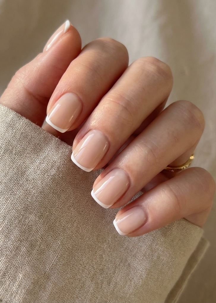 A close-up of a hand showing short square nude nails with crisp white French tips, paired with a beige linen sleeve.