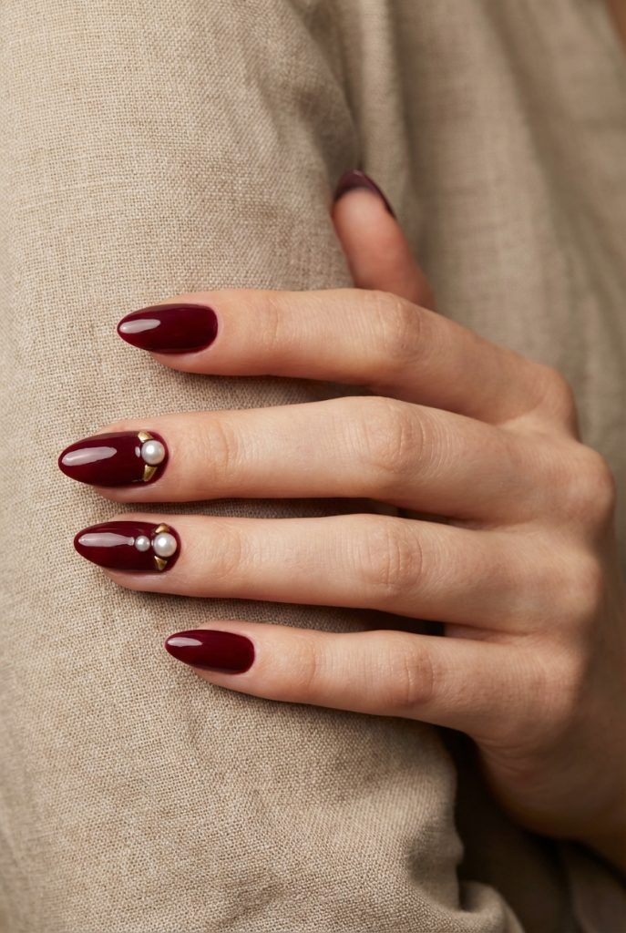 Close-up of almond-shaped burgundy nails with pearl and gold half-moon designs on two fingers—illustrating a moody pearl nail art idea.