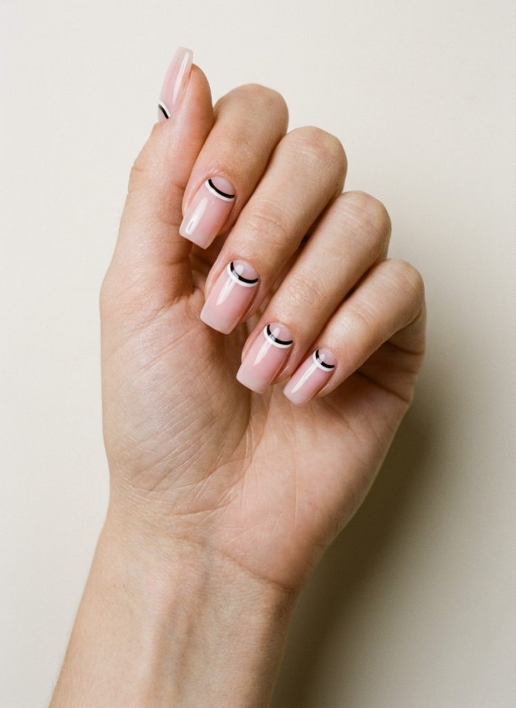 Close-up of a hand with squared nails painted in blush pink, detailed with bold black semi-circles near the cuticle for a playful reverse French nail art idea.