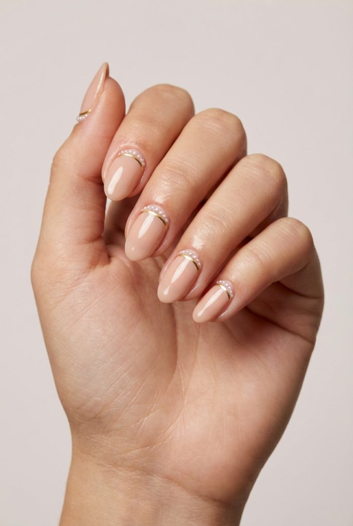 Hand with soft nude nails, highlighting a pearl nail art idea that features tiny pearls and gold lines forming graceful curves at the base of short almond-shaped nails.