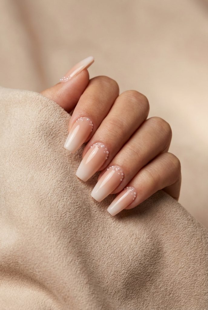 A woman's hand with square-shaped nude ombré nails featuring a pearl nail art idea—tiny pearls forming an elegant crescent along each cuticle, captured in soft lighting.