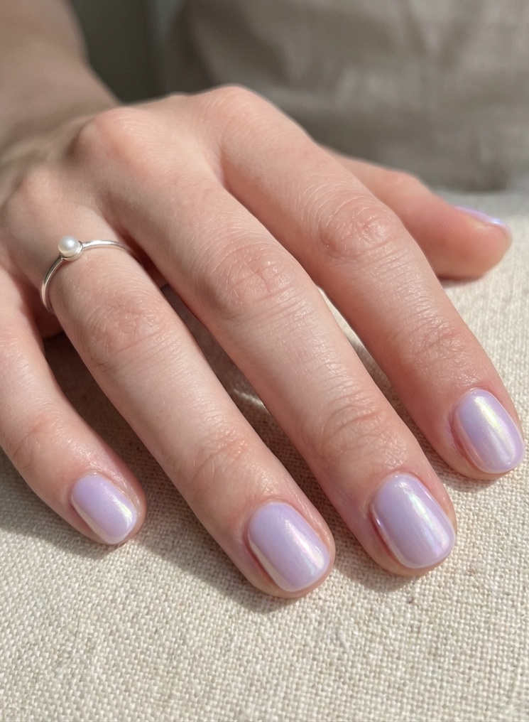 Feminine hand resting on fabric, showing a glazed nail art idea in pastel lilac with a glowing, pearly finish across short natural nails.