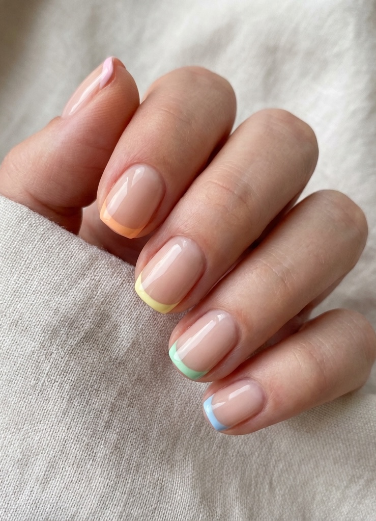 Short nail art idea featuring a hand with short nude nails and pastel French tips in soft pink, yellow, mint green, and light blue.