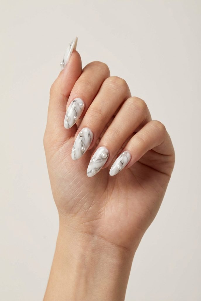 A hand with almond-shaped nails featuring white and gray marble polish, adorned with clusters of pearls, illustrating a pearl nail art idea that mirrors moonlight on stone.