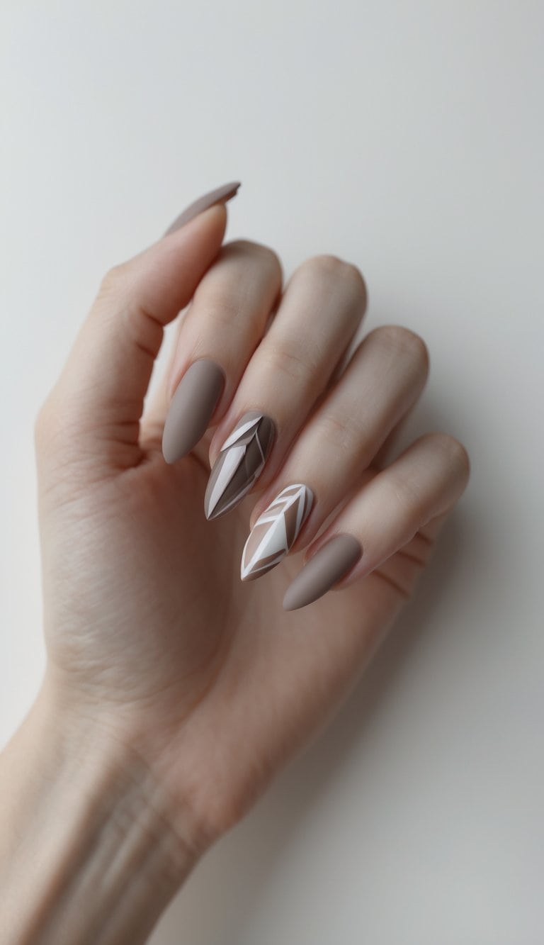 A hand is shown with matte taupe nails and two accent nails featuring sharp white geometric lines, creating a modern graduation nail art idea.