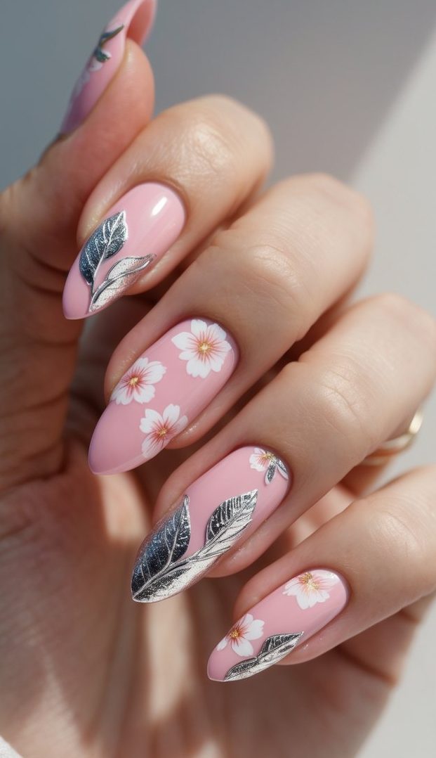 Seen from an angled perspective, a hand displays long almond nails styled in a pink and silver nail art idea, featuring pale pink polish contrasted by white blossoms and silver leaf motifs.