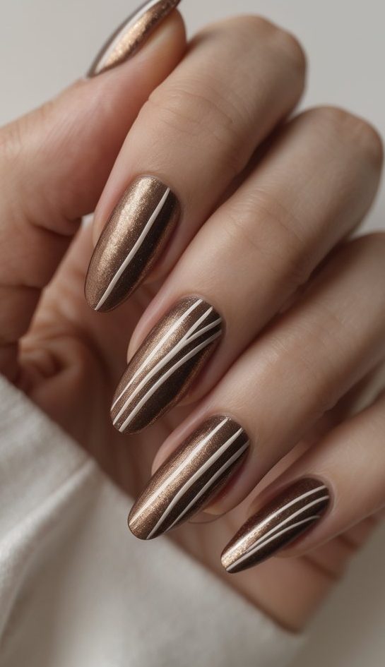 close-up of a hand with bronze shimmer nails, some adorned with fine white stripes, showcasing a minimal shimmer nail art idea