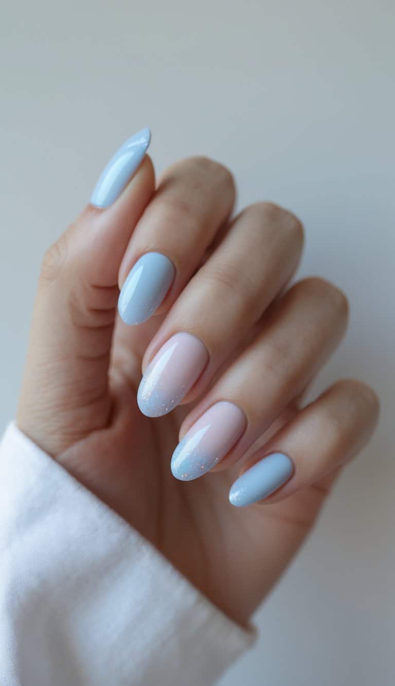 A hand shown up close with almond-shaped nails painted in light blue and pale pink shades, featuring a faint glitter sheen concentrated near the nail tips.