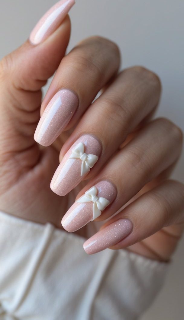 Blush-toned glitter nails with sculpted white bows, designed to resemble romantic gift wrapping in a dreamy shimmery nail art idea.