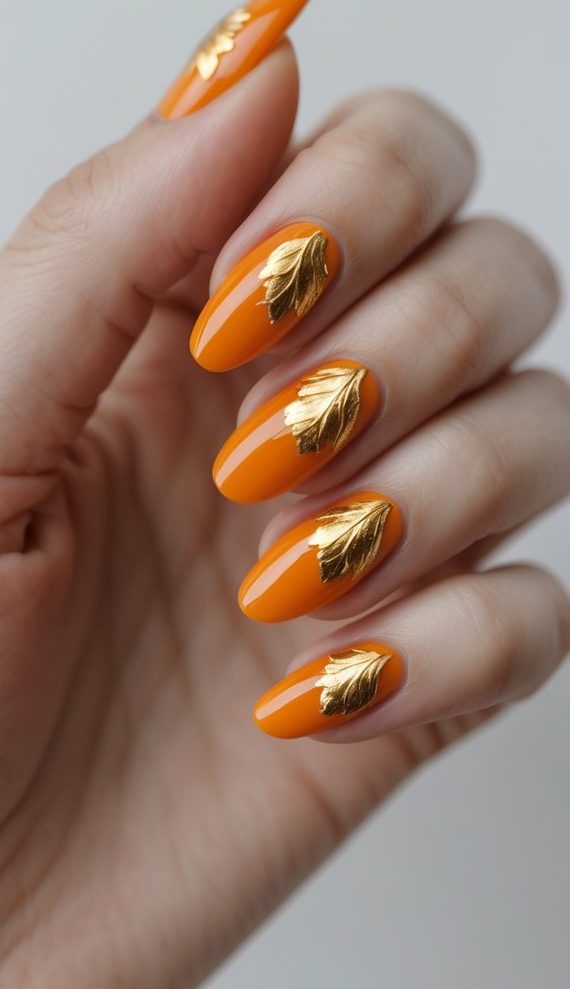 Hand showing glossy orange almond-shaped nails with 3D golden leaf accents on each nail, highlighting a bold glossy nail art idea perfect for fall.