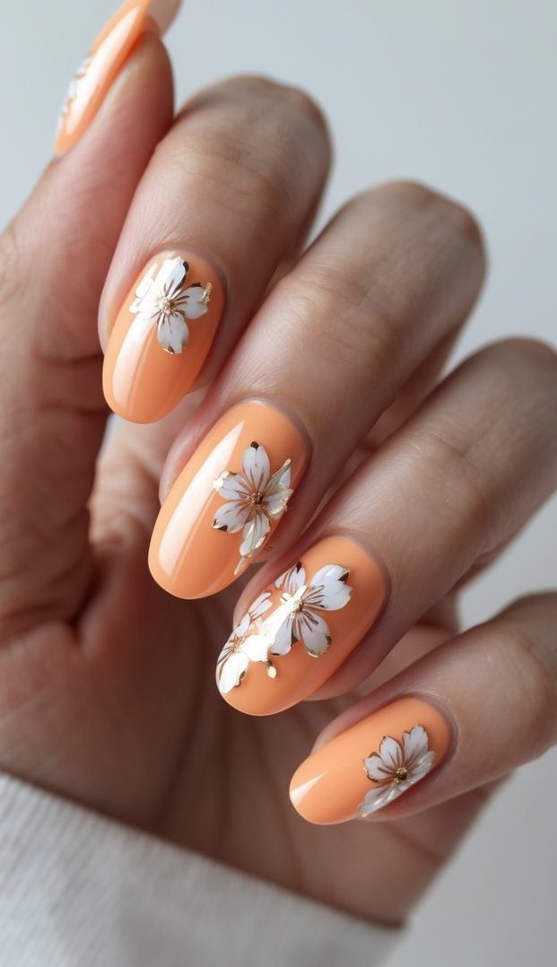Hand with oval nails painted in apricot orange and adorned with white floral designs outlined in gold, showing a poetic glossy nail art idea.