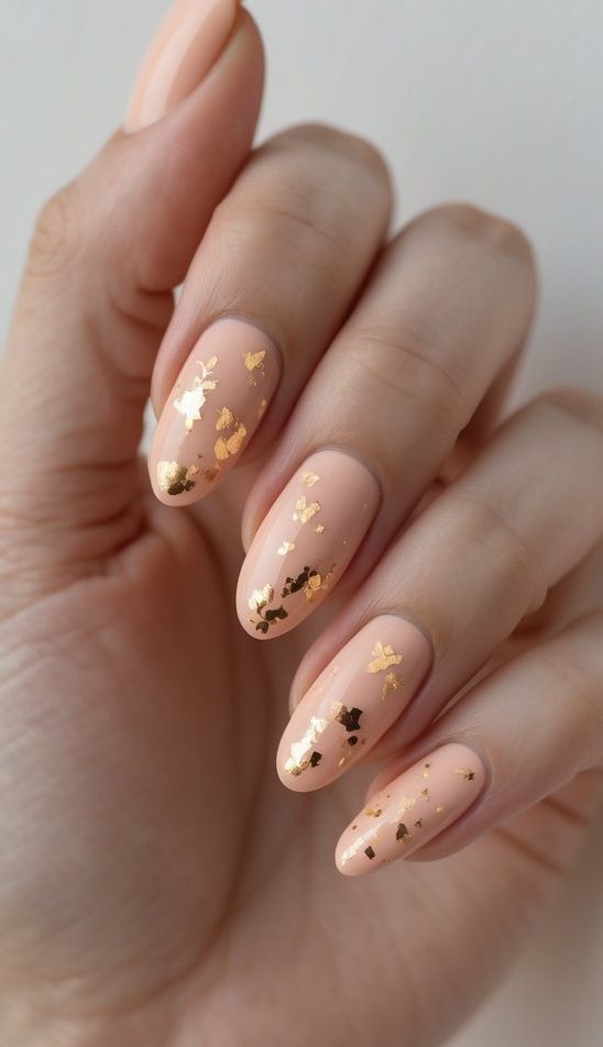 Close-up of a hand featuring a foil nail art idea with nude-peach polish and fun scattered gold foil resembling party confetti.