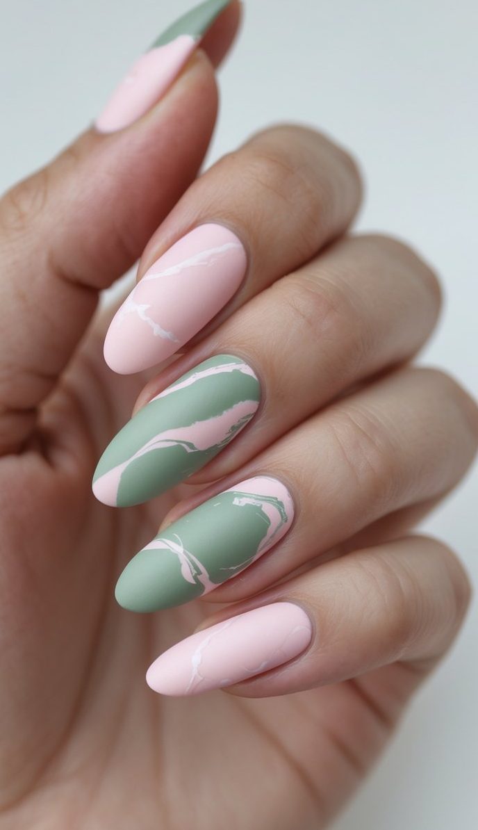 Soft-focused hand with almond-shaped nails painted in alternating matte sage green and blush pink, featuring a marble nail art idea with delicate white veining details