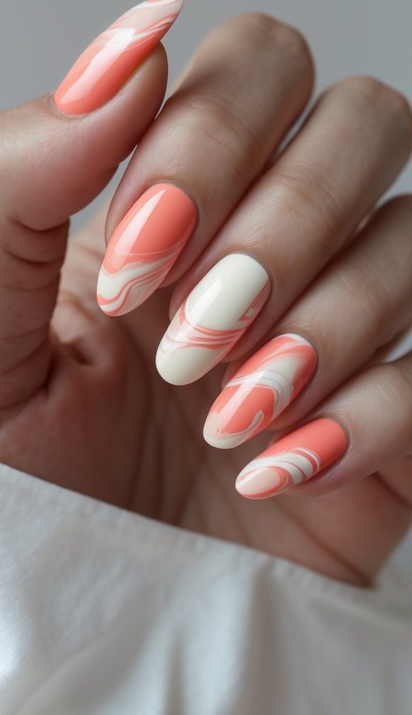 Hand with glossy coral and ivory nails styled in a marble nail art idea, combining gentle swirls and color-blocking in warm, summery tones