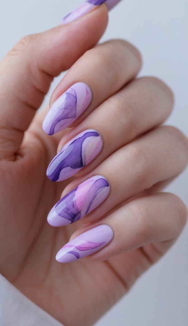 A close-up of a hand with almond-shaped nails painted in soft marbled patterns of lavender, purple, and pink—showcasing a refined watercolor nail art idea.