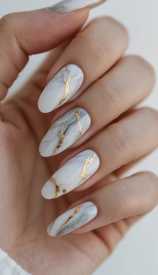 A polished hand shows white nails etched with gray marbling and jagged gold foil, resembling stone—an elevated watercolor nail art idea.