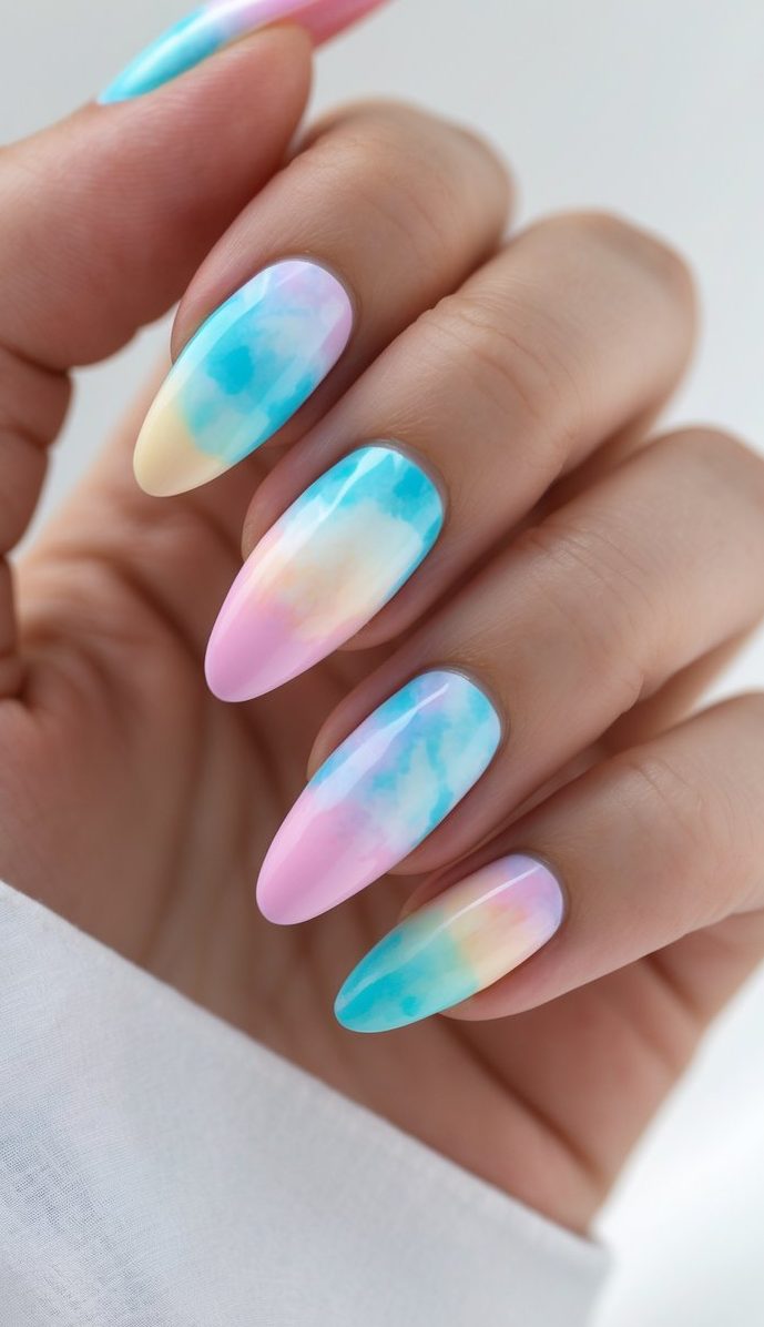 A woman's hand shows a tie dye nail art idea using pastel pink, baby blue, and soft yellow, resembling gentle watercolors on almond-shaped nails.