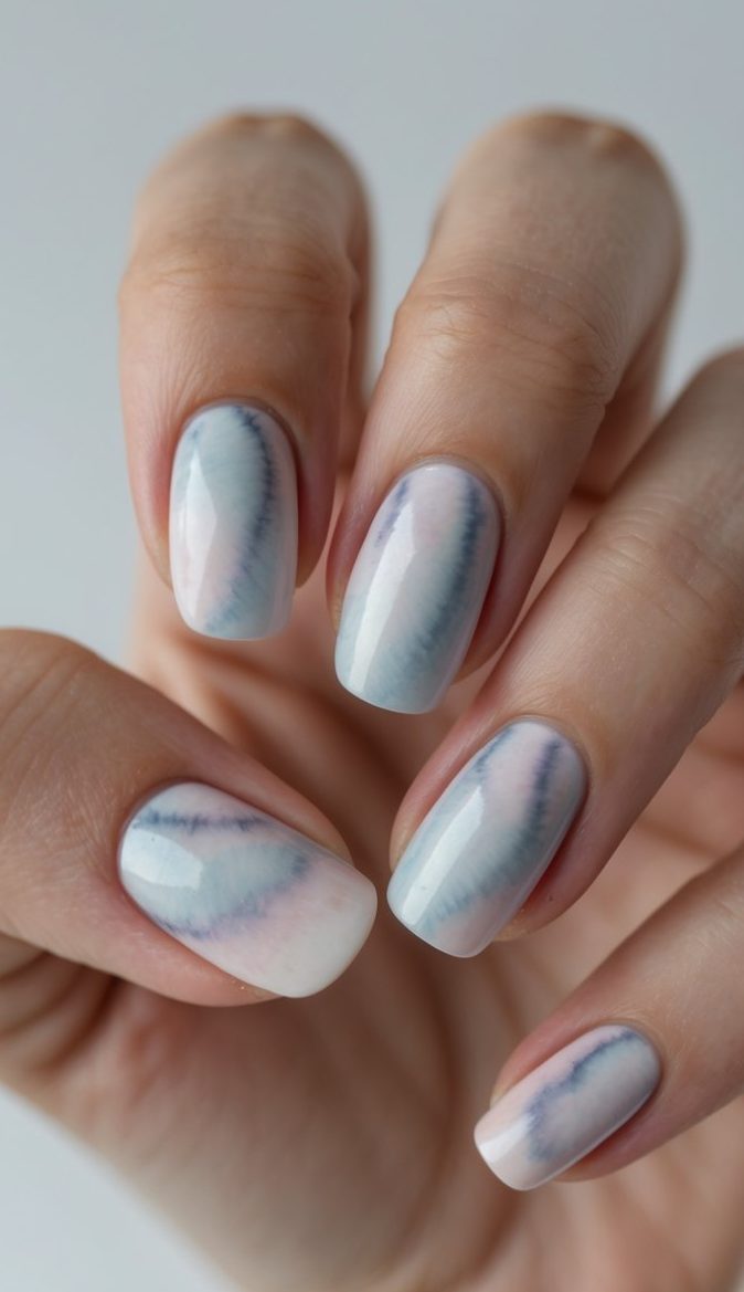 A hand with short rounded nails shows a tie dye nail art idea using hazy blends of blue, lilac, and blush in a dreamy watercolor style.