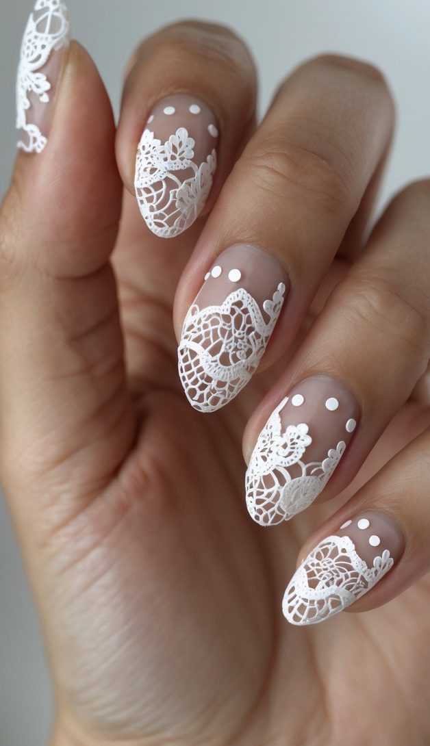 Almond nails painted in a soft nude-pink base with dainty white lace overlays and dotted details, mimicking a vintage lace nail art idea.