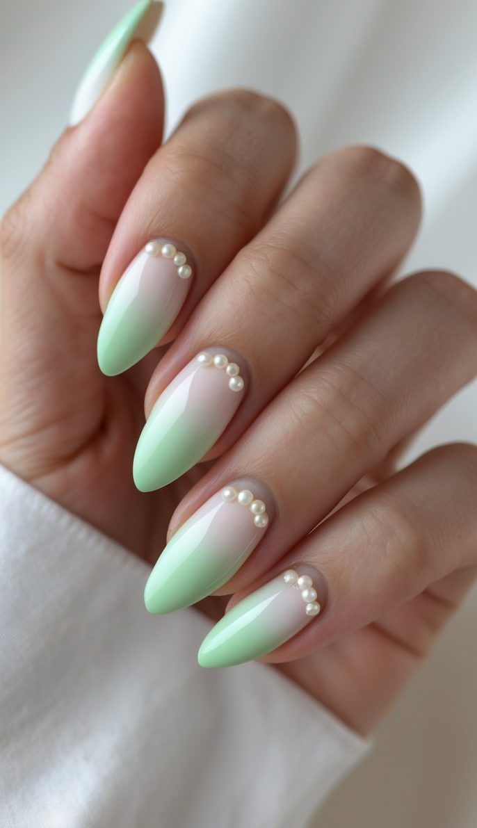 Almond-shaped nails in a coquette nail art idea with glossy mint green tips and pearl studs along a soft pink translucent base on a hand.