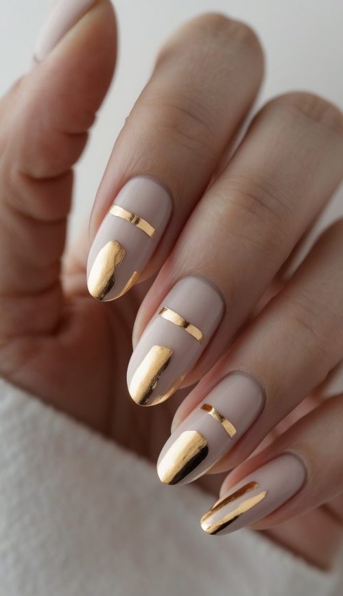 Close-up of a hand with nude nails styled in a minimalist design, featuring geometric gold foil accents and band-like detailing—a sleek sophisticated nail art idea.