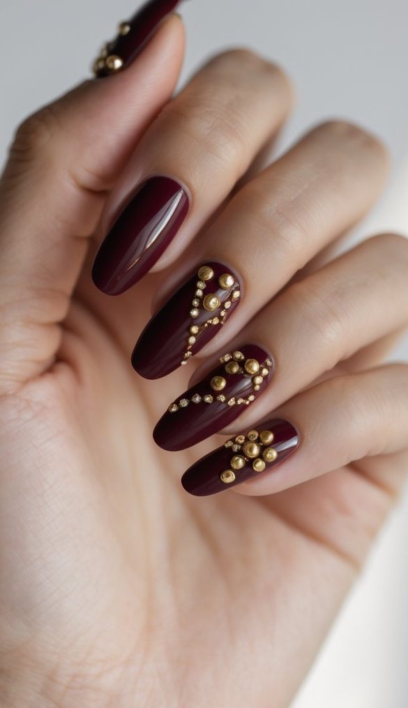 Burgundy nails detailed with layered gold studs and pearls in geometric patterns—this sophisticated nail art idea delivers bold texture and metallic drama.
