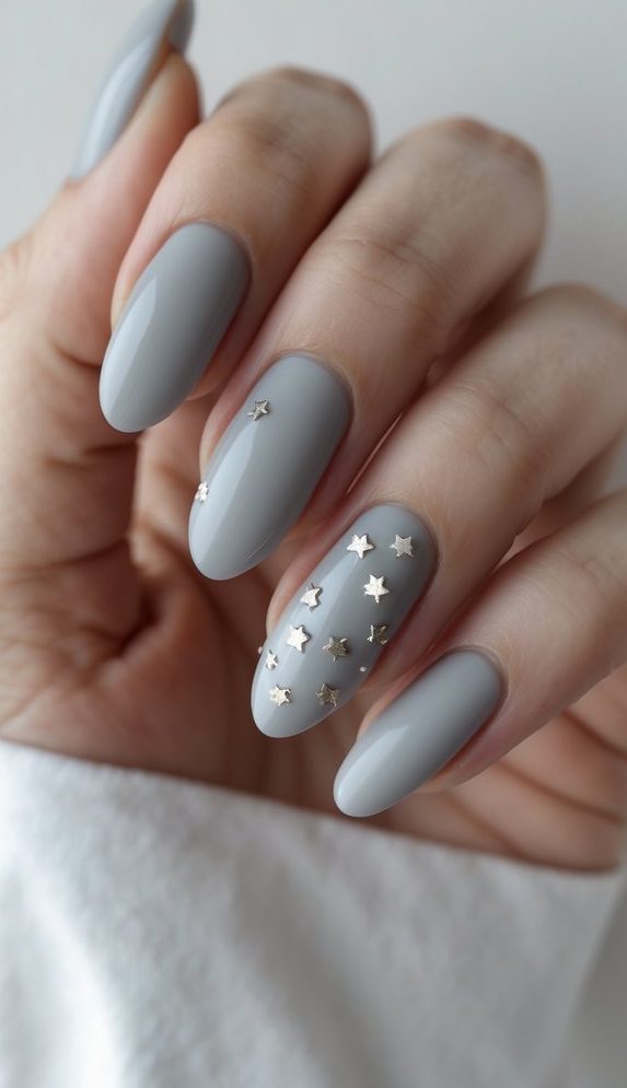 A hand showing pale grey almond-shaped nails with small silver stars on accent nails, highlighting a delicate nail art idea with a celestial touch.