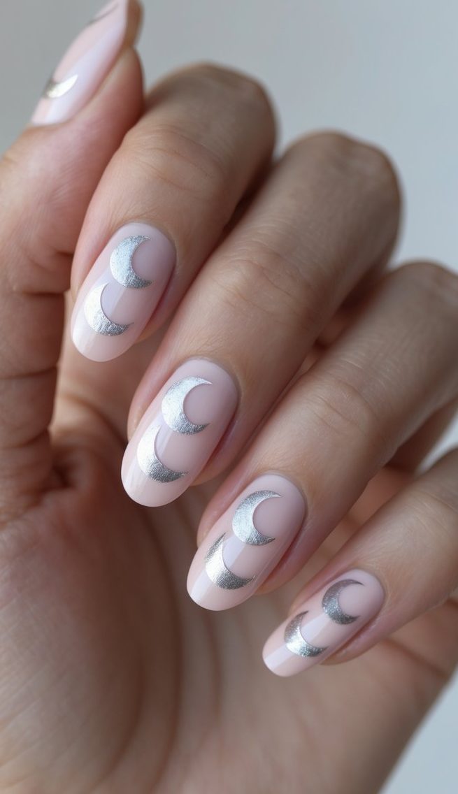A hand displays nude nails adorned with metallic silver crescent moons—an ethereal and delicate nail art idea inspired by the quiet glow of moonlight.