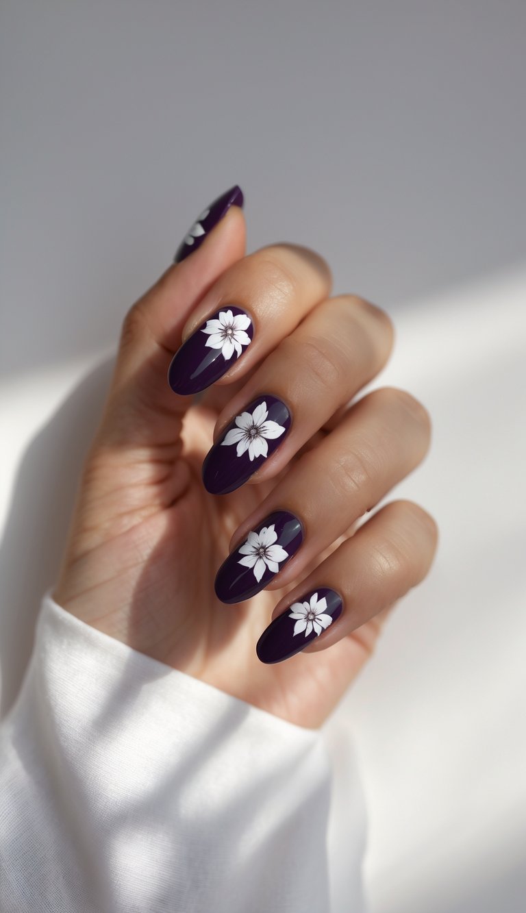 A relaxed hand pose reveals a purple and white nail art idea, showcasing dark purple nails enhanced with hand-painted white floral designs against a neutral background.