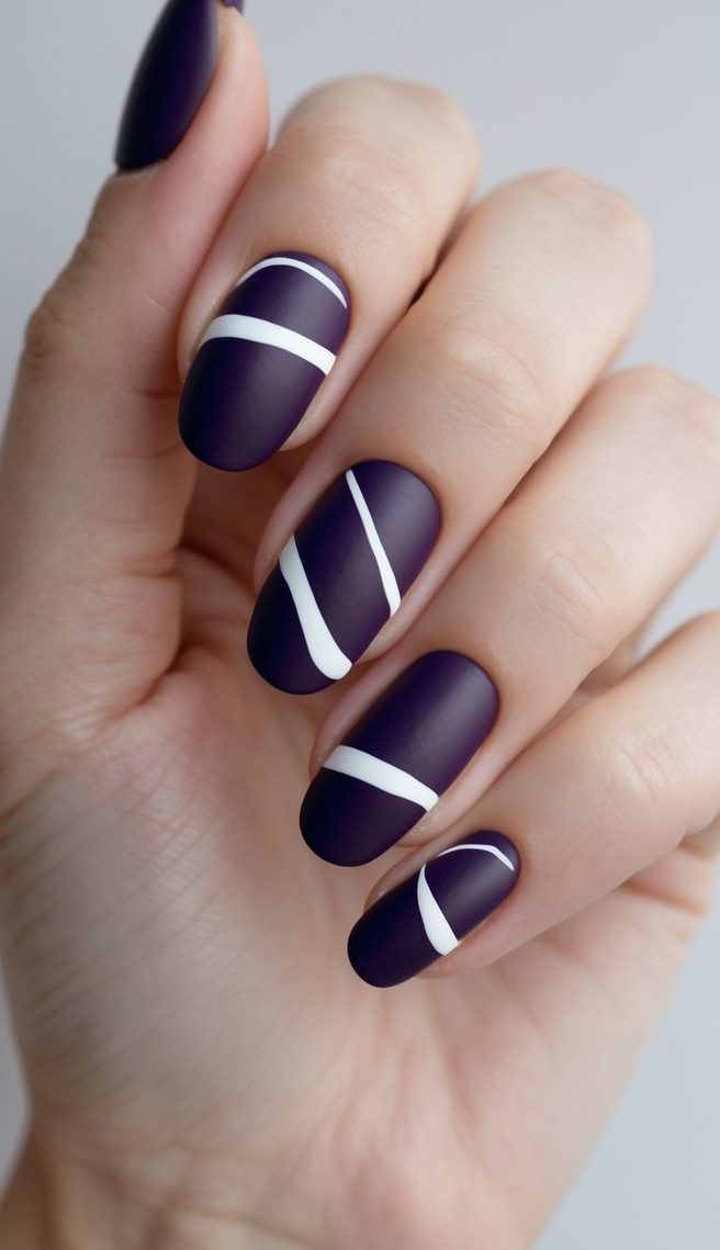 Fingers shown up close wearing matte plum nails with white curved stripe details, presenting a bold and refined purple and white nail art idea.
