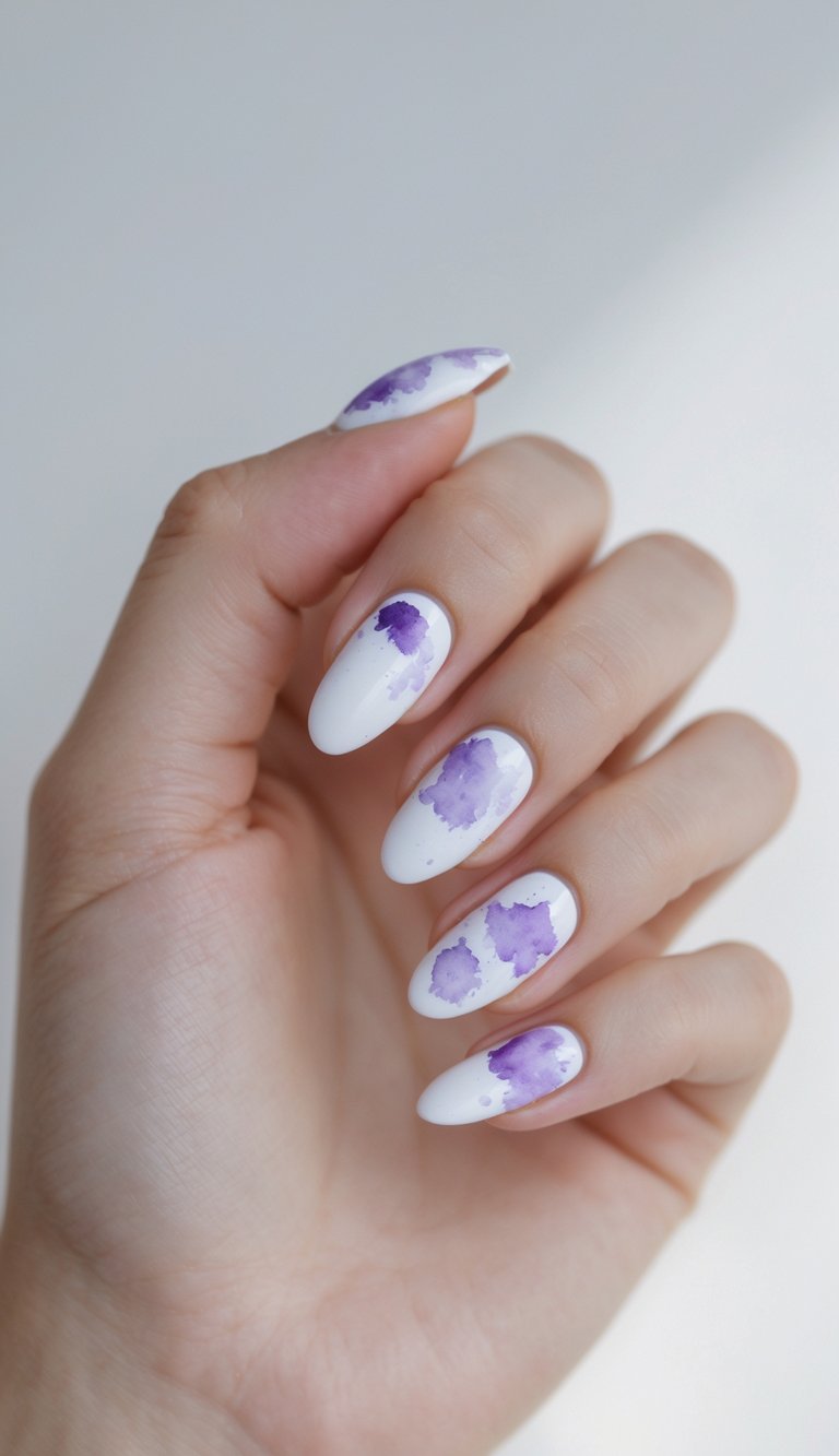 A close-up of a hand shows white almond-shaped nails decorated with soft purple watercolor-style blotches, creating an airy purple nail art idea with a painted effect.
