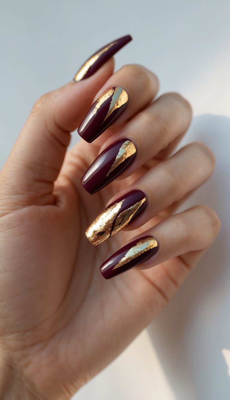 Close-up of one hand showing burgundy coffin-shaped nails with gold foil accents on a white background.